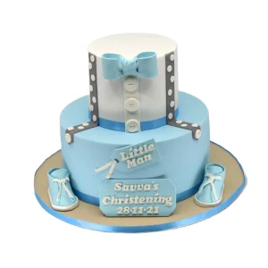 Two-tier christening cake for a baby boy featuring a shirt-style top tier with blue bow tie, white buttons, and polka-dot suspenders. Bottom tier is light blue with a ribbon, fondant tags reading "Little Man" and "Savva's Christening 28-11-21." Decorated with fondant baby shoes on a gold board with blue trim.