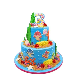 Two-tier underwater-themed birthday cake in vibrant blue, decorated with fondant sea creatures, coral, starfish, and Baby Shark characters. Features a polka-dot number 3 topper and a speech bubble with the name “ARA!” for a third birthday celebration.