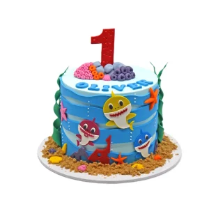 Ocean-themed first birthday cake featuring blue fondant waves, colorful Baby Shark characters, sea creatures, coral, and a red number 1 topper. Name “OLIVER” displayed on top.