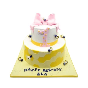 Two-tier bee-themed birthday cake with honeycomb texture, edible bees, and a pink bow topper. A sweet and playful centrepiece for a first birthday celebration.