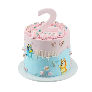 Two-tone second birthday cake with pink and blue frosting, topped with pink swirls, sprinkles, a large number 2, and Bluey and Bingo characters. Name “Mya” written on the side.