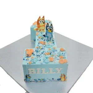 Number-shaped first birthday cake in light blue frosting with orange and blue swirls, colorful sprinkles, and Bluey and Bingo toppers. Name “BILLY” displayed on the front.