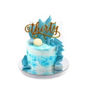 Blue and white marbled 30th birthday cake topped with gold “thirty” script, blue macarons, and candy shards.