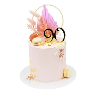 Elegant 90th birthday cake by Amarantos Cakes featuring blush pink textured buttercream, gold leaf accents, peach and cream macarons, and a luxe dried floral topper with black and gold '90' centerpiece.