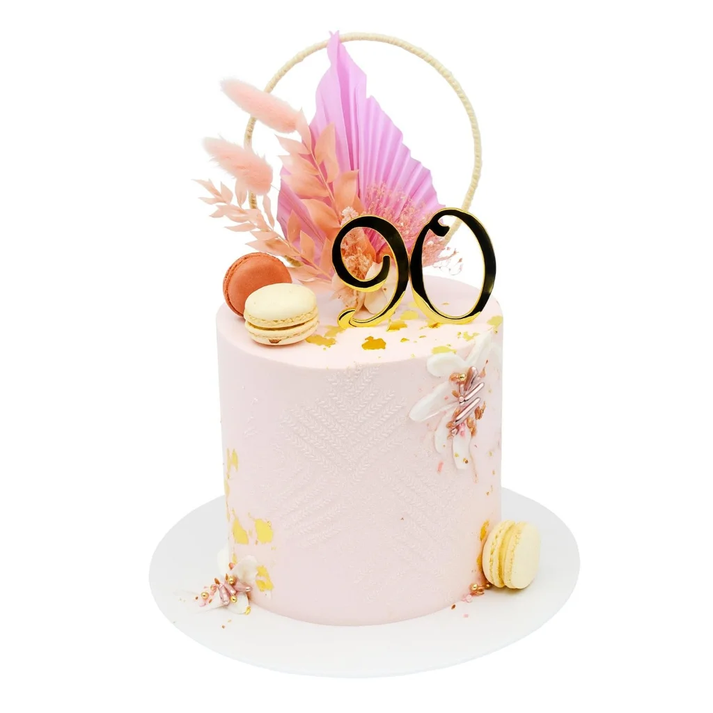 Elegant 90th birthday cake by Amarantos Cakes featuring blush pink textured buttercream, gold leaf accents, peach and cream macarons, and a luxe dried floral topper with black and gold '90' centerpiece.