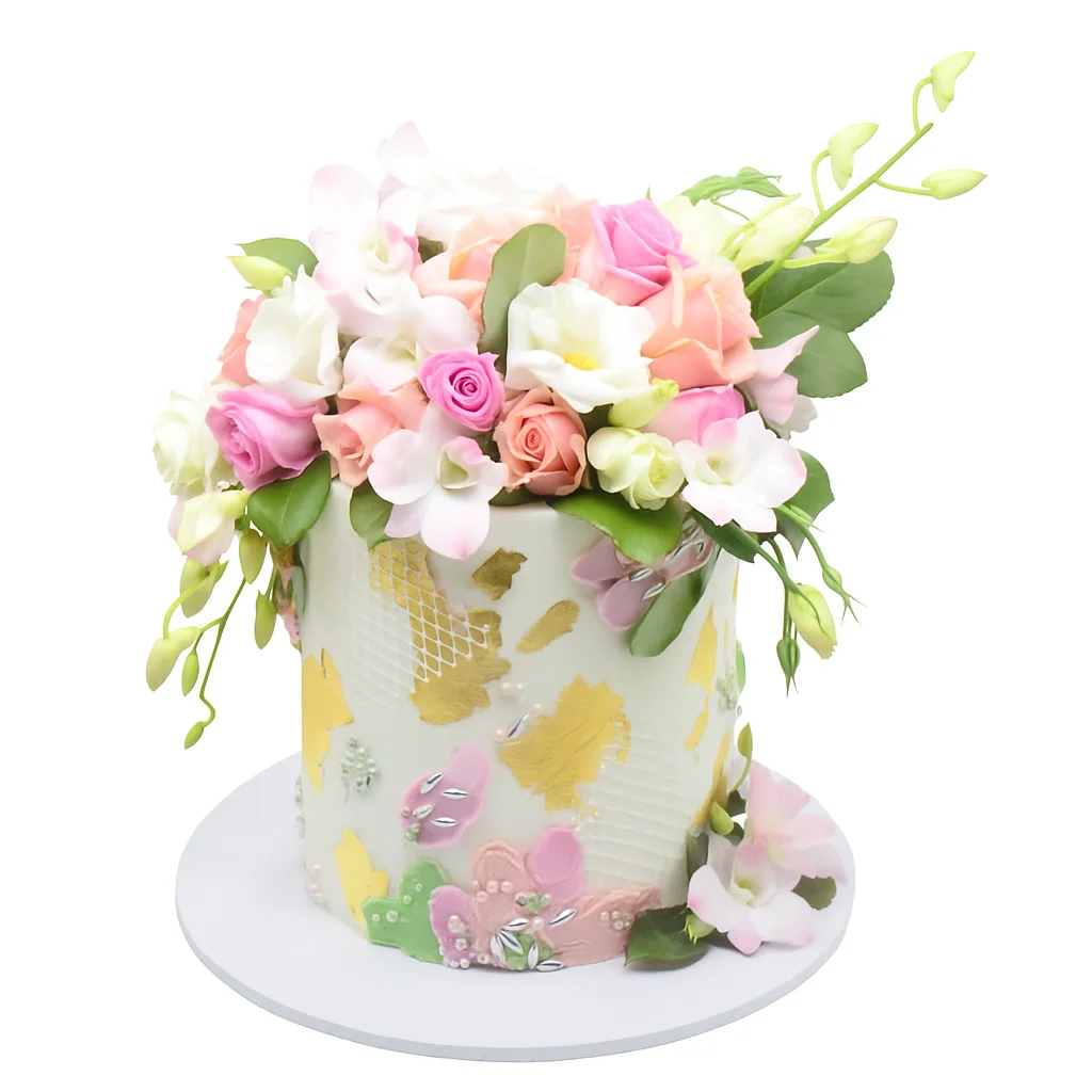 Elegant floral celebration cake by Amarantos Cakes featuring white textured buttercream, gold leaf accents, pastel side florals, and a lush bouquet of pink, peach, and white flowers with greenery.