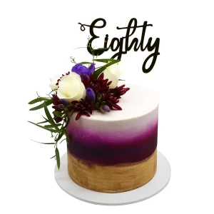 Elegant 80th birthday cake by Amarantos Cakes featuring a smooth gold-to-purple gradient buttercream, fresh white roses, purple and dark red blooms, green foliage, and a cursive “Eighty” topper.