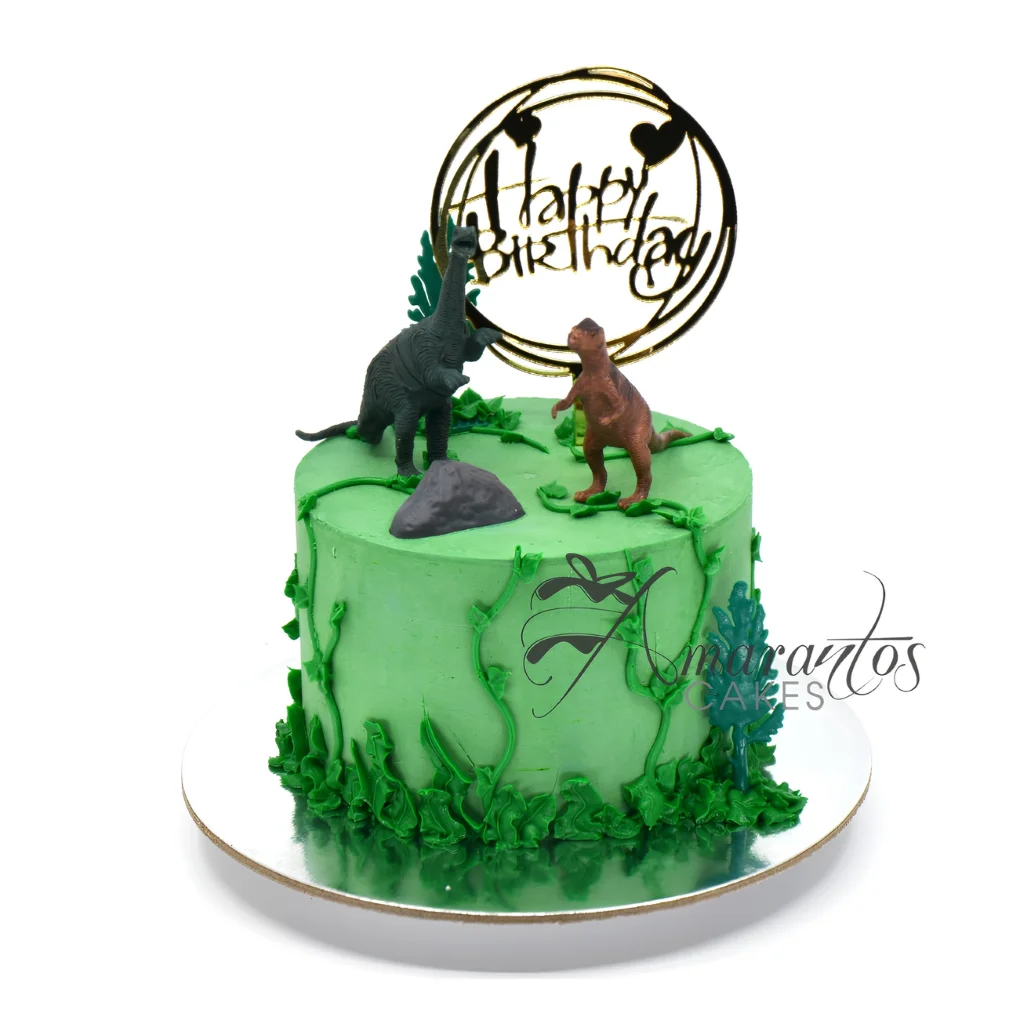 Dinosaur-themed birthday cake by Amarantos Cakes featuring green buttercream, icing vines and foliage, plastic dinosaur figurines, a grey rock decoration, and a stylised “Happy Birthday” topper with heart accents.