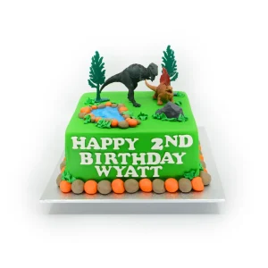Dinosaur birthday cake with green fondant, dino figurines, pond, stones, trees, and jungle foliage.