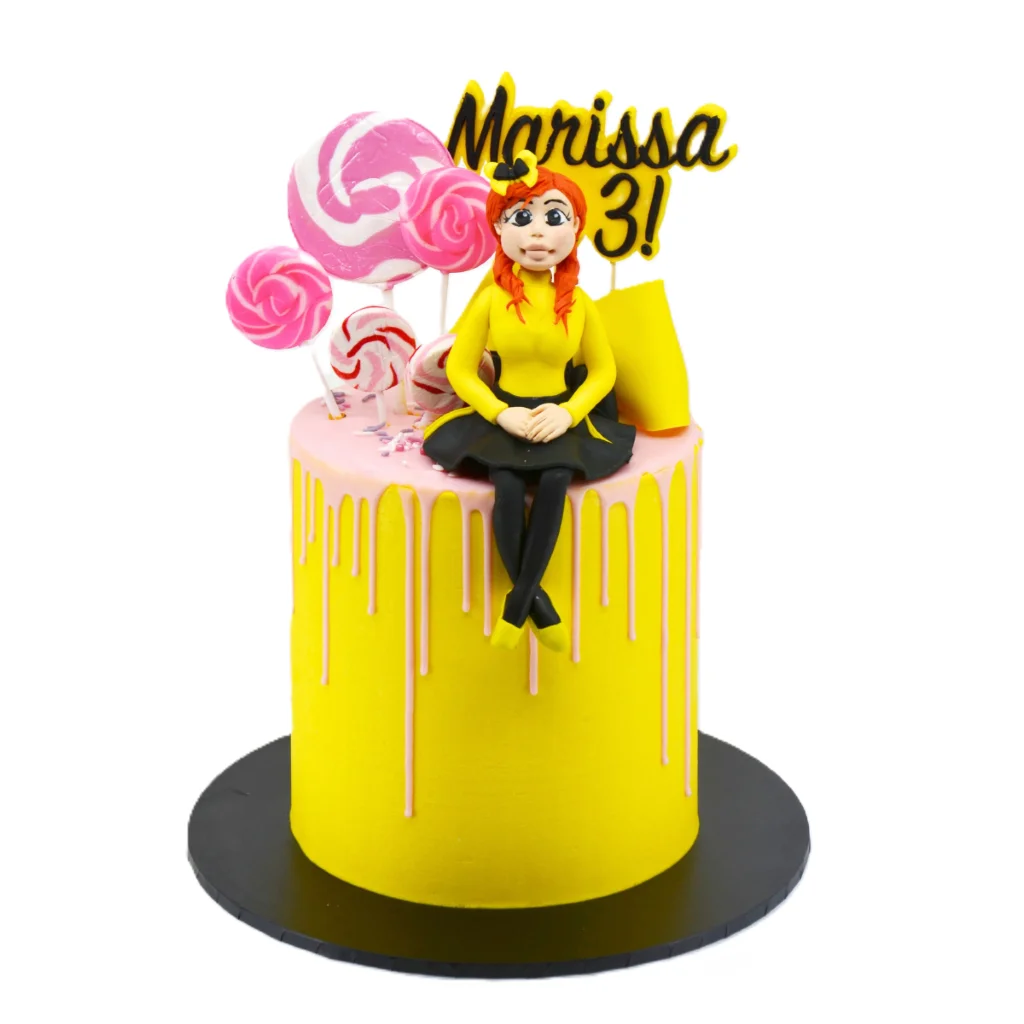 Bright and playful third birthday cake by Amarantos Cakes featuring yellow buttercream, pink drip icing, Emma from the Wiggles with red hair, decorative lollipops, a yellow party hat, and a “Marissa 3!” topper.