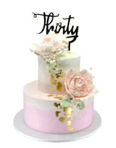 A two‑tier pink and white birthday cake decorated with gold leaf, soft pink and white sugar flowers, and a gold “Thirty” topper, displayed on a white cake board