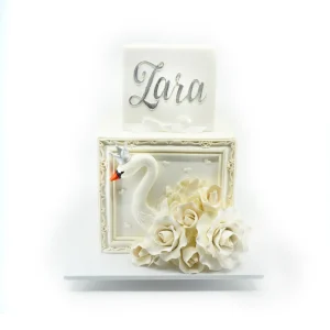 White two-tier square cake with silver name topper, sculpted swan with crown, ornate frame detailing, and fondant roses.
