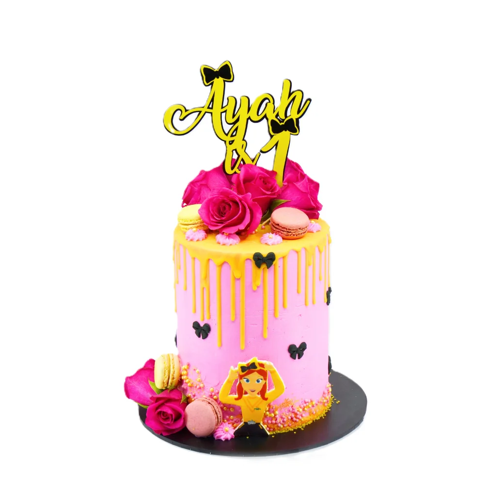 Pink and yellow drip birthday cake with black bows, roses, colorful macarons, cartoon girl topper, and “Ayan is 1” sign.